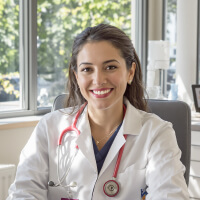 A doctor in a white coat with a stethoscope around her neck smiles while seated in an office with windows in the background.