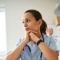 A person wearing medical scrubs and a stethoscope sits thoughtfully with hands clasped under their chin, looking to the side.