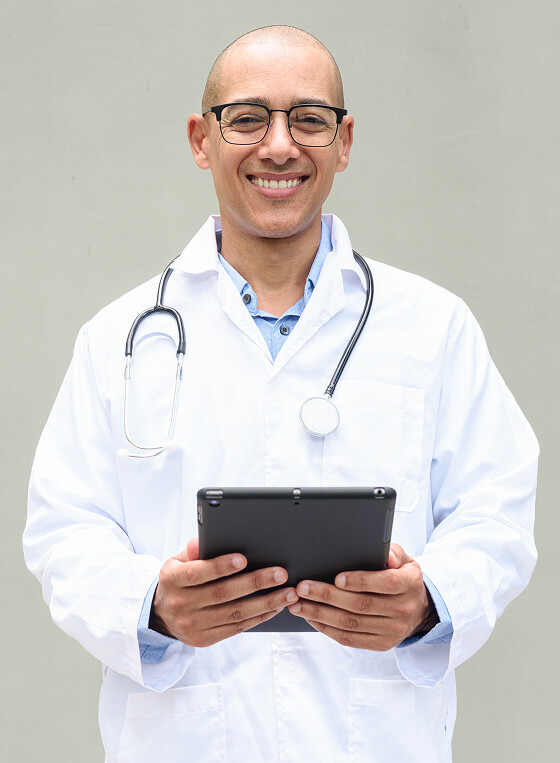A doctor wearing a white coat and stethoscope holds a tablet and smiles at the camera against a plain background.