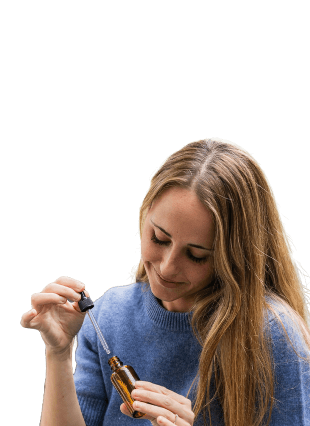 Woman with long hair in a blue sweater holds a dropper and small amber bottle, dispensing liquid with a neutral expression against a plain light background.