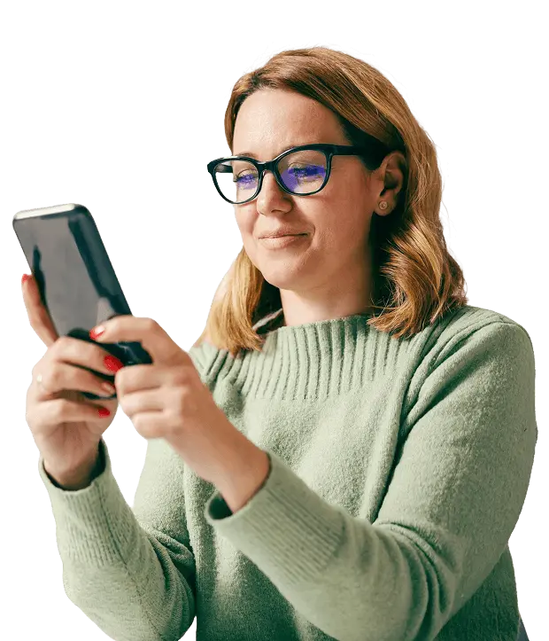 A woman wearing glasses and a green sweater holds and uses a smartphone, looking at the screen with a focused expression.