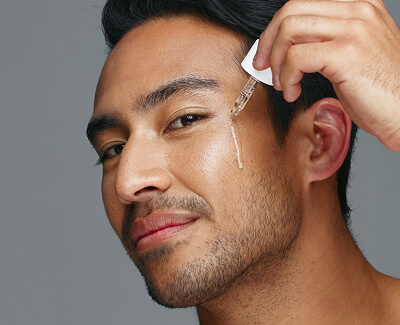 A man applies a skincare serum to his face using a dropper against a plain gray background.