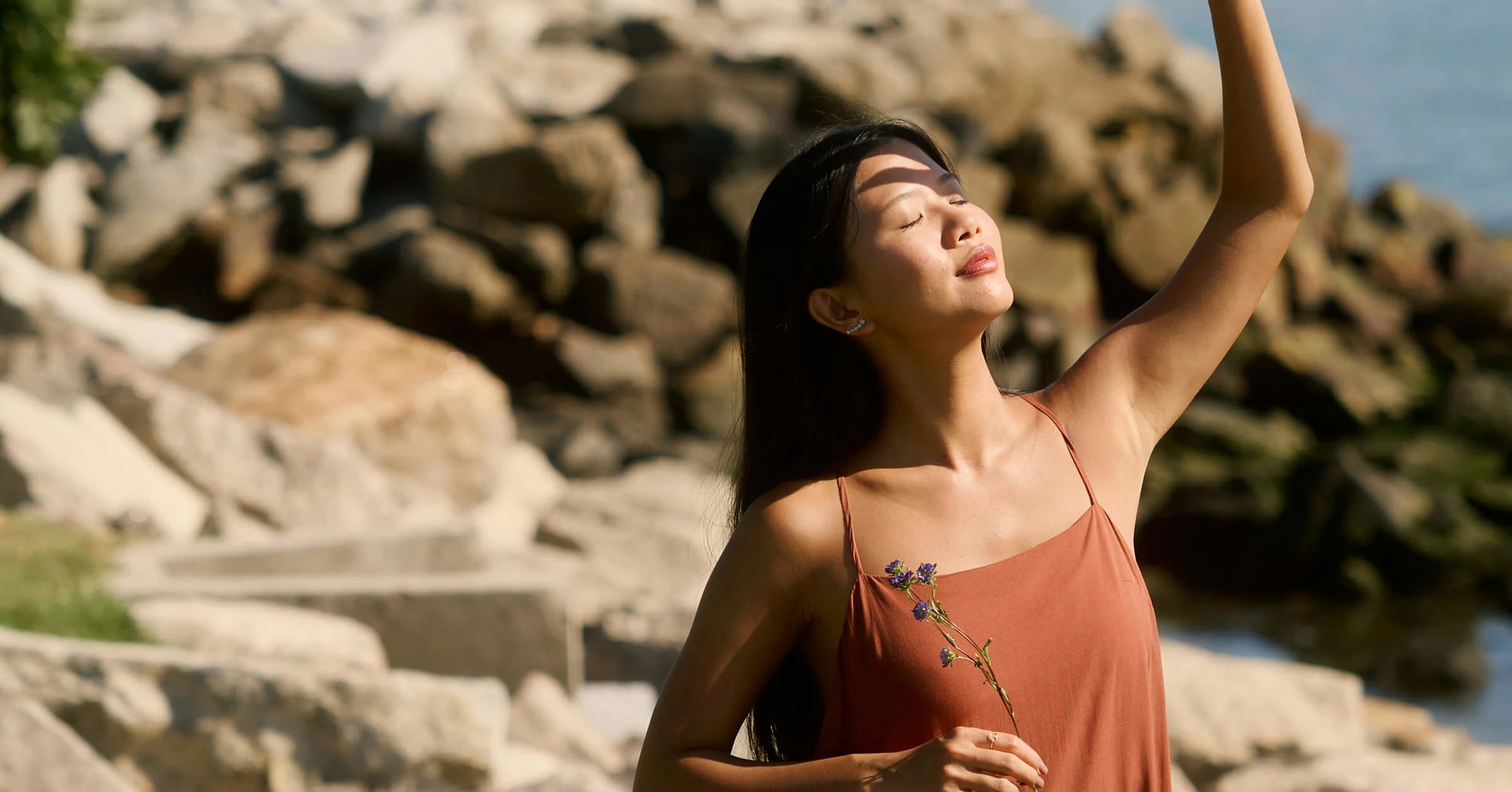 A woman in a sleeveless dress stands outdoors holding a small flower, eyes closed, and raises her arm toward sunlight near rocky terrain.