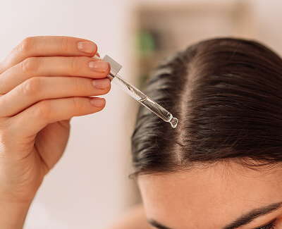 A person applies liquid to their scalp using a dropper, focusing on the part in their hair.