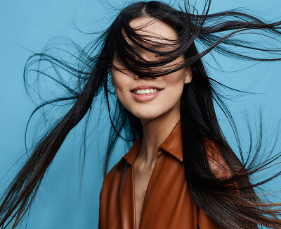 Woman with long dark hair smiling as her hair blows across her face, wearing a brown top and standing against a blue background.