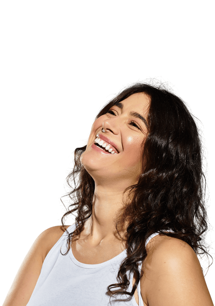 Woman with long dark curly hair, wearing a white tank top, smiling and laughing while looking upward against a plain light background.