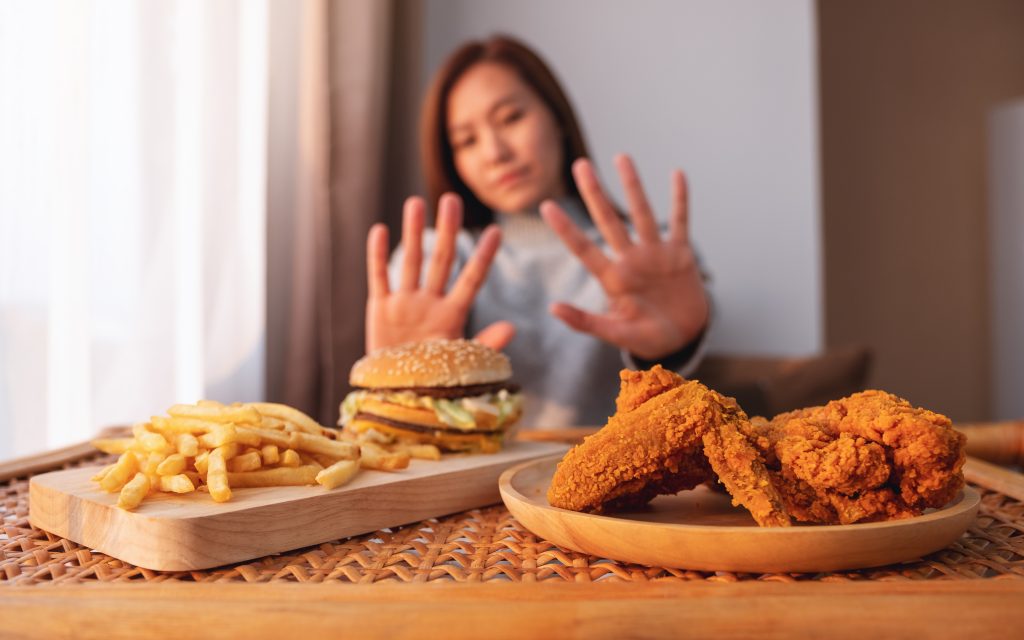 A person with their hands up, eagerly responding to hunger signals, gesturing at a wooden table laden with a burger, fries, and fried chicken.