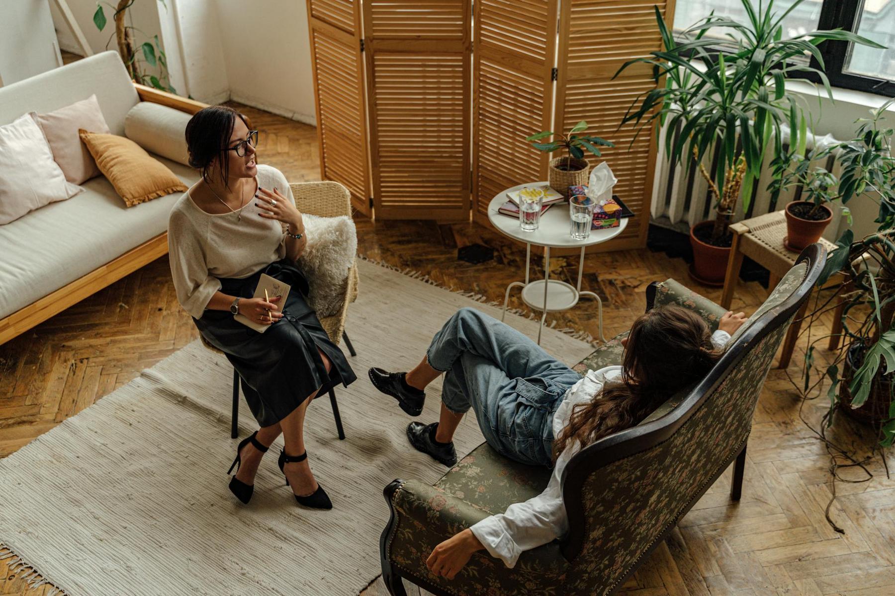 Two women are sitting and talking in a cozy living room with plants, a rug, and a round table with books and glasses. One woman sits upright with a notebook; the other reclines on a sofa.