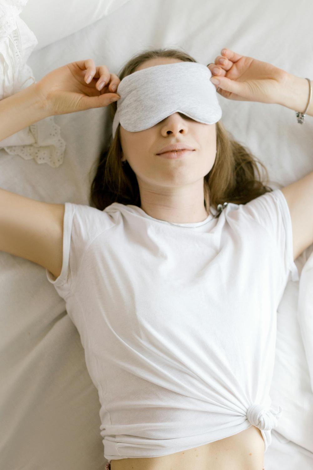 A person lying on a bed wearing a white T-shirt adjusts a light gray sleep mask over their eyes.