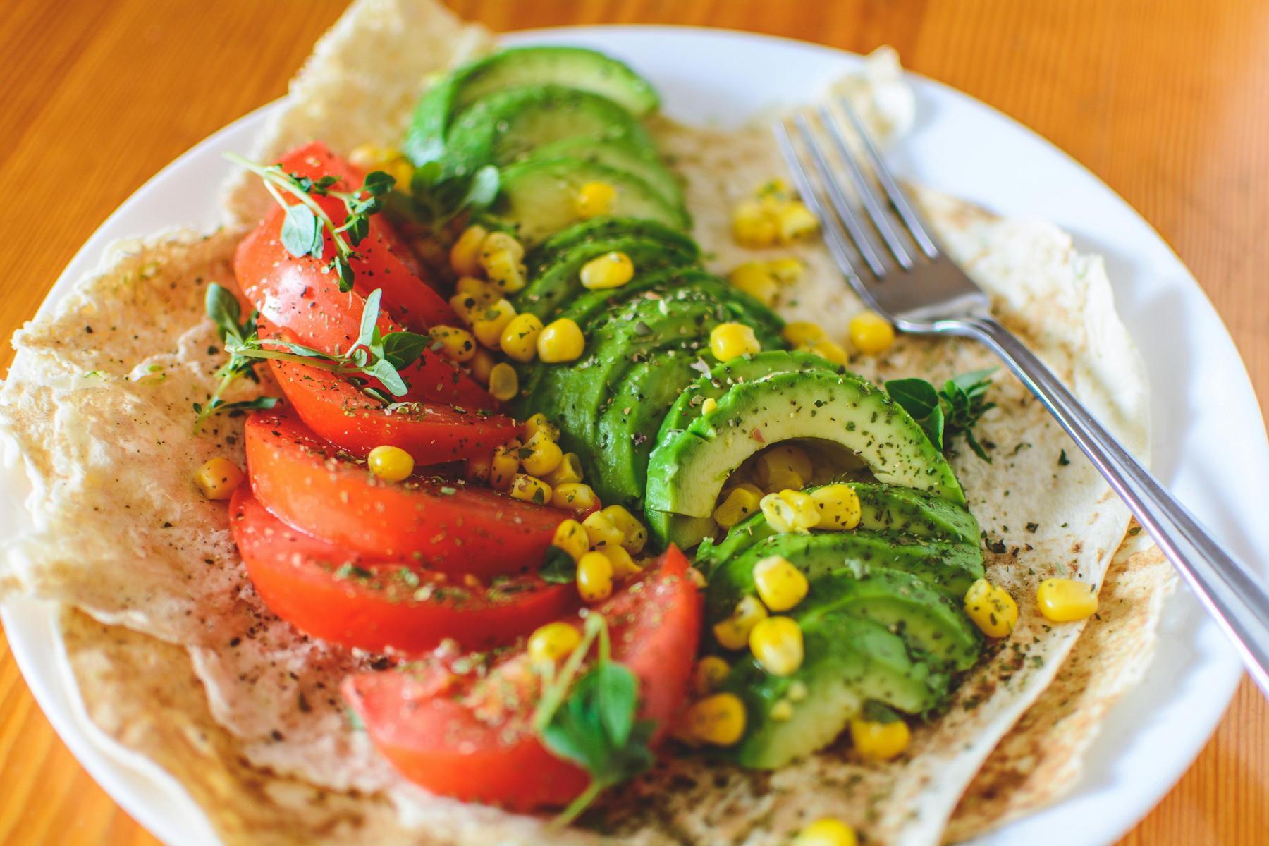 A plate with sliced tomatoes, avocado, sweet corn, fresh herbs, and spices on flatbread, with a fork on the side.