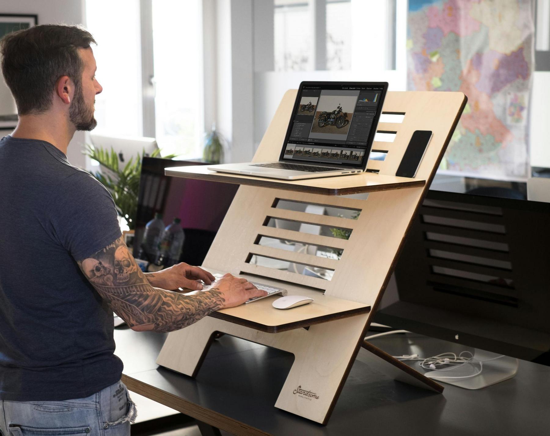 A man stands at a modern wooden standing desk with a laptop and mouse on separate shelves in a contemporary office setting.