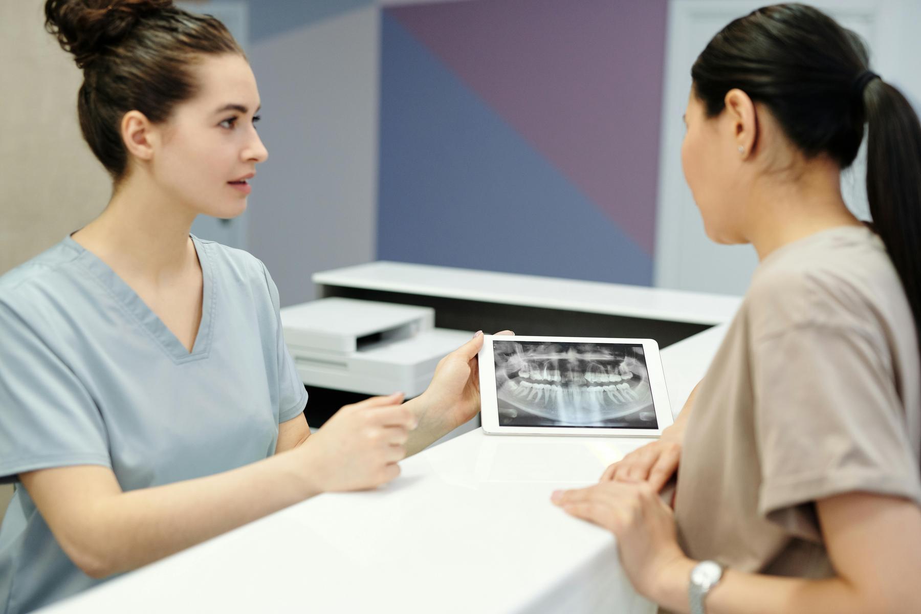 Two healthcare professionals in scrubs discuss a dental X-ray displayed on a tablet at a reception counter.
