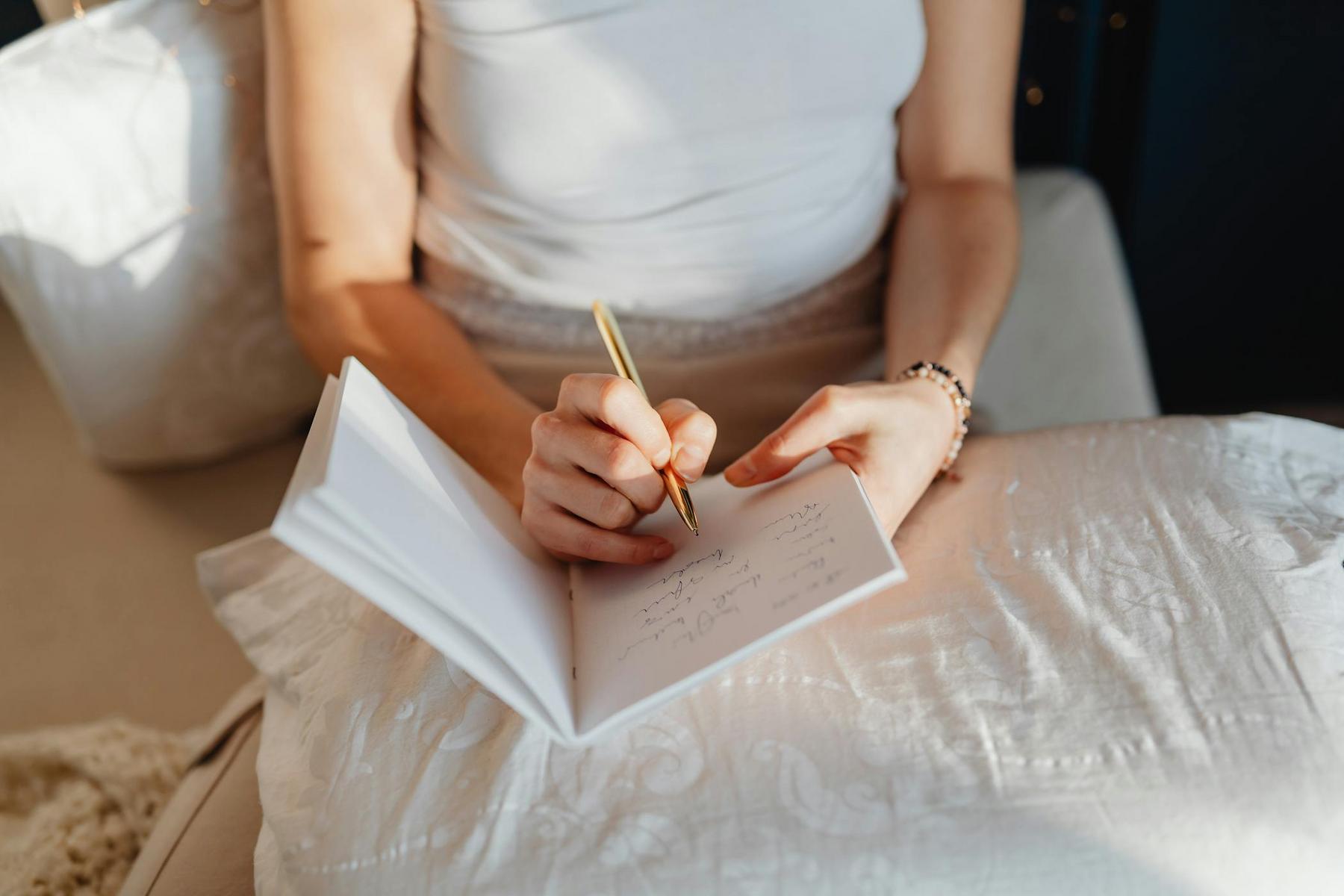 A person sits holding a notebook and pen, writing on a page while resting their arm on a white pillow.