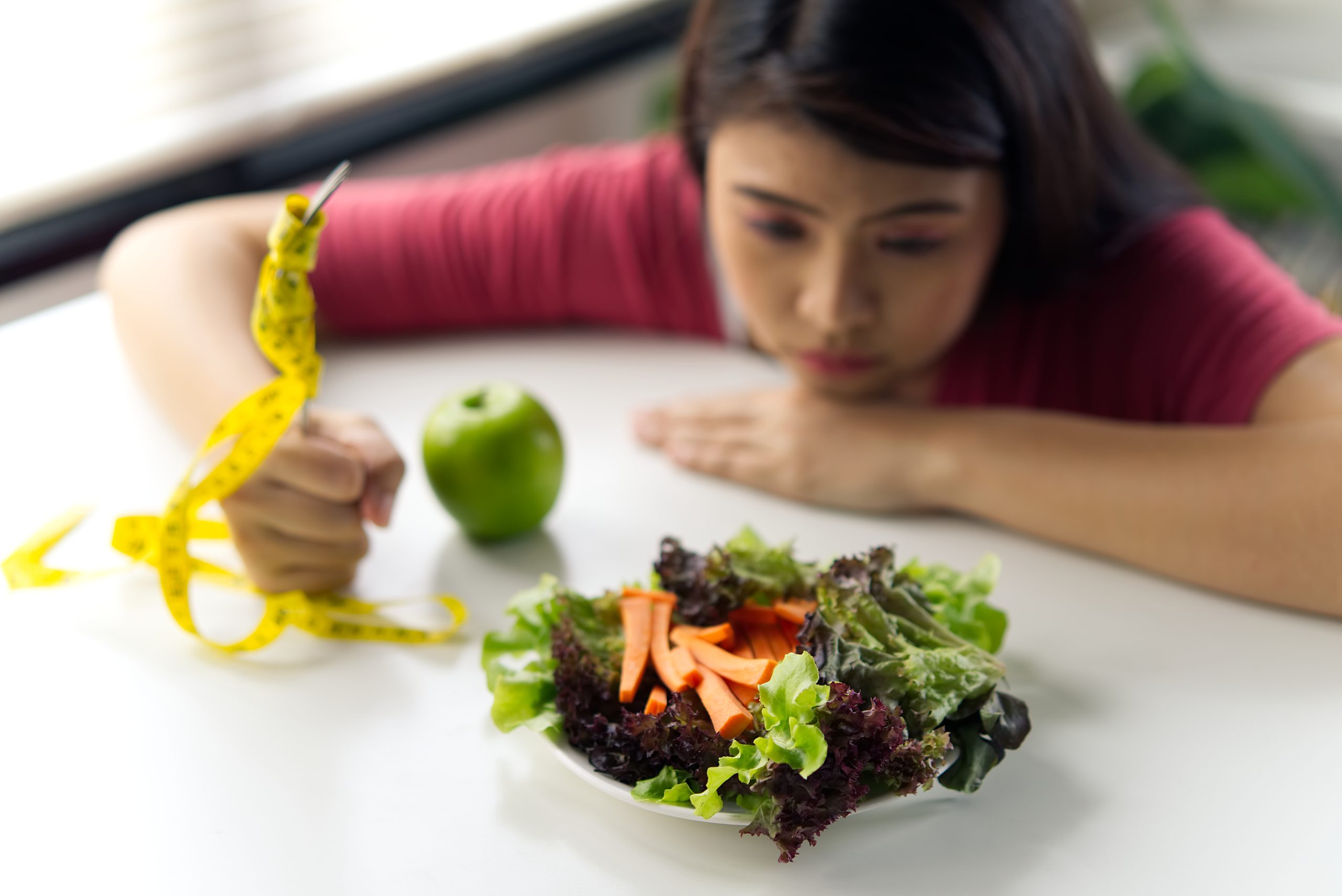 A woman examines a salad and an apple on the table, holding a yellow measuring tape in her hand, as she contemplates their connection to weight loss goals.