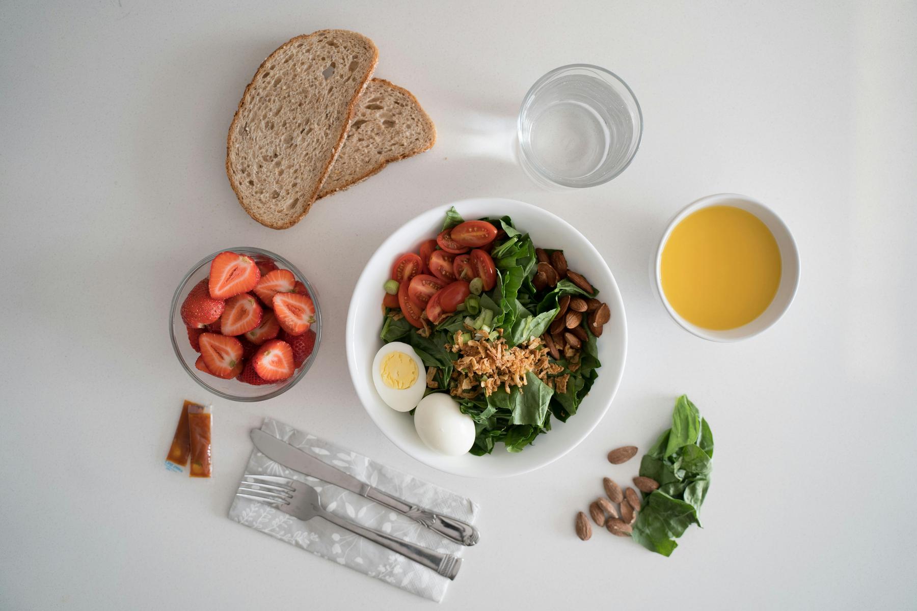 A meal with a salad bowl, boiled egg, almonds, sliced tomatoes, greens, two bread slices, a glass of water, orange juice, strawberry bowl, cutlery on a napkin, and dressing packets.