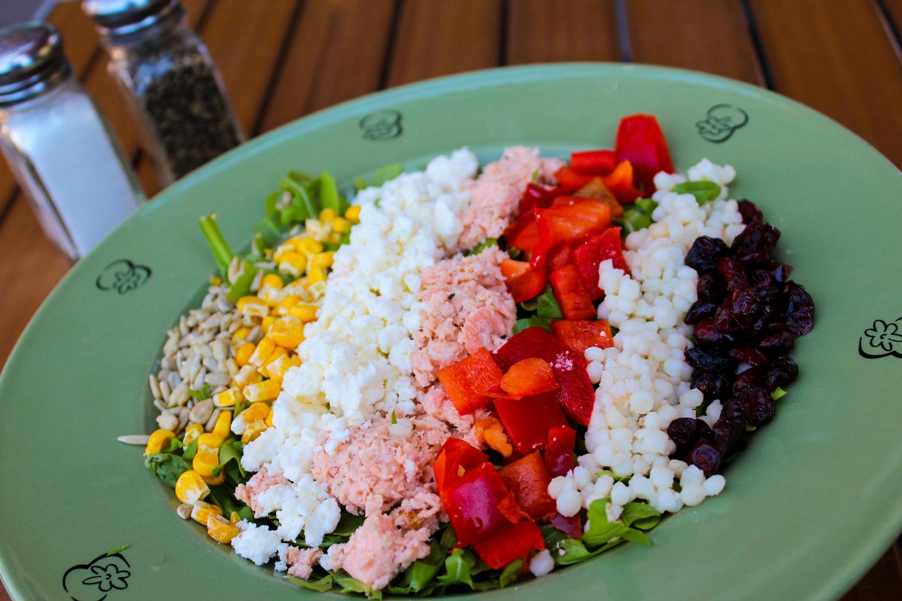 A salad on a green plate featuring corn, sunflower seeds, feta cheese, tuna, red peppers, couscous, and dried cranberries, arranged in sections. Salt and pepper shakers are in the background.