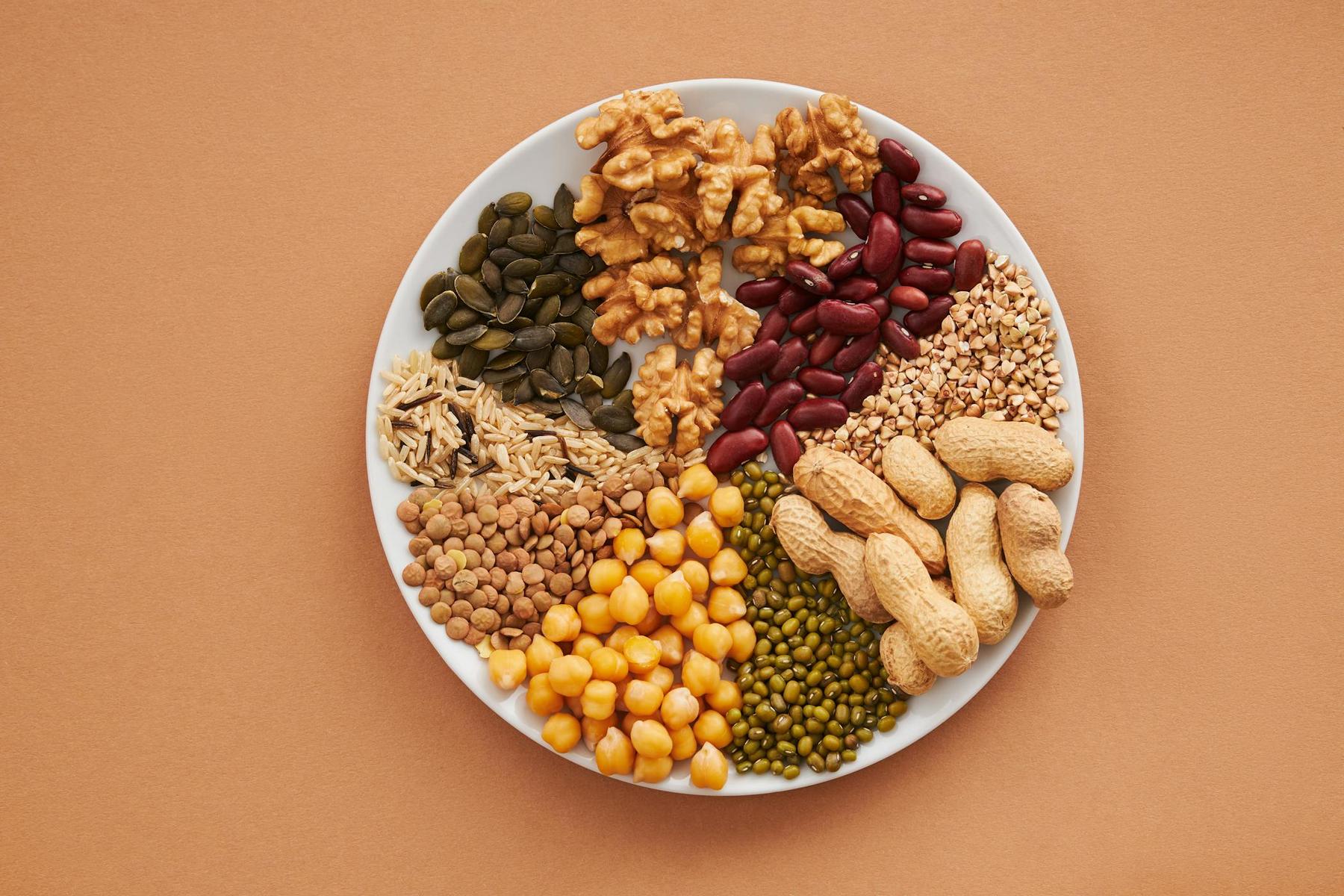 A white plate with walnuts, kidney beans, pumpkin seeds, lentils, chickpeas, mung beans, peanuts, and sunflower seeds on a brown surface.