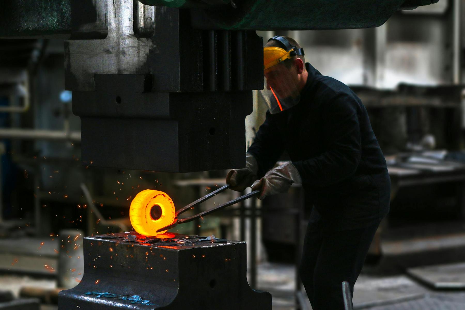 A worker wearing protective gear uses tongs to handle a glowing hot metal piece under a large industrial press, with sparks flying.