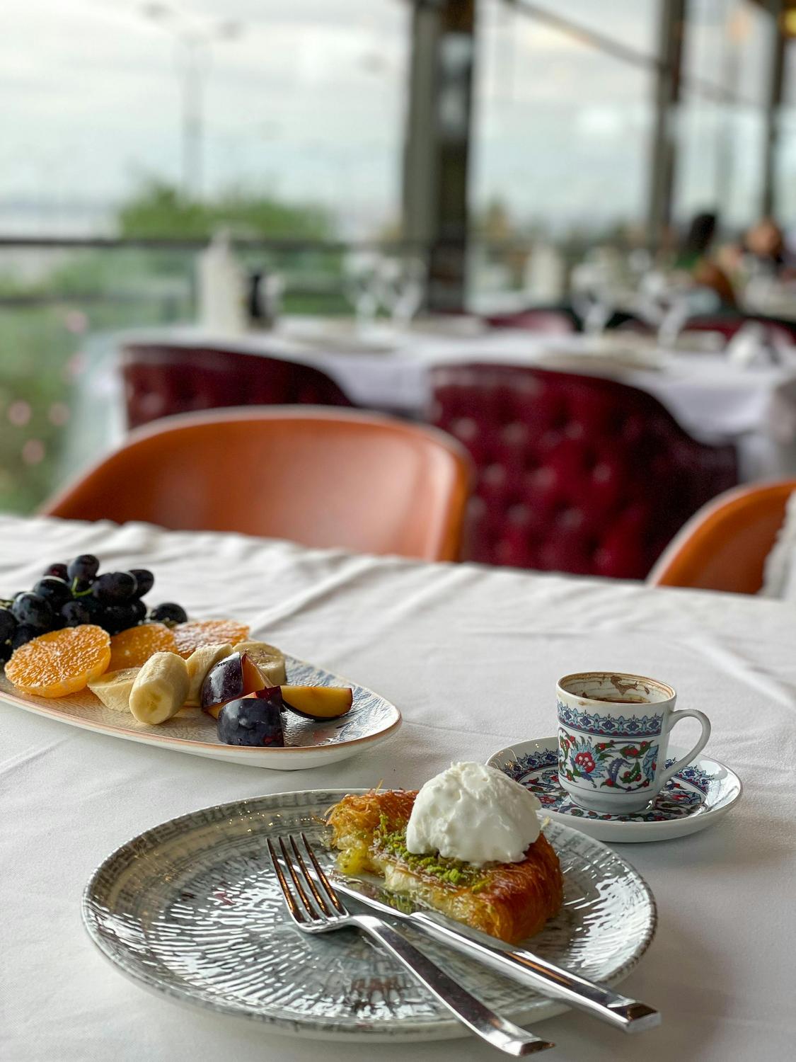 A table set with a plate of assorted fresh fruit, a dessert with cream, a fork and knife, and a cup of coffee in a decorated cup, in a restaurant with large windows.