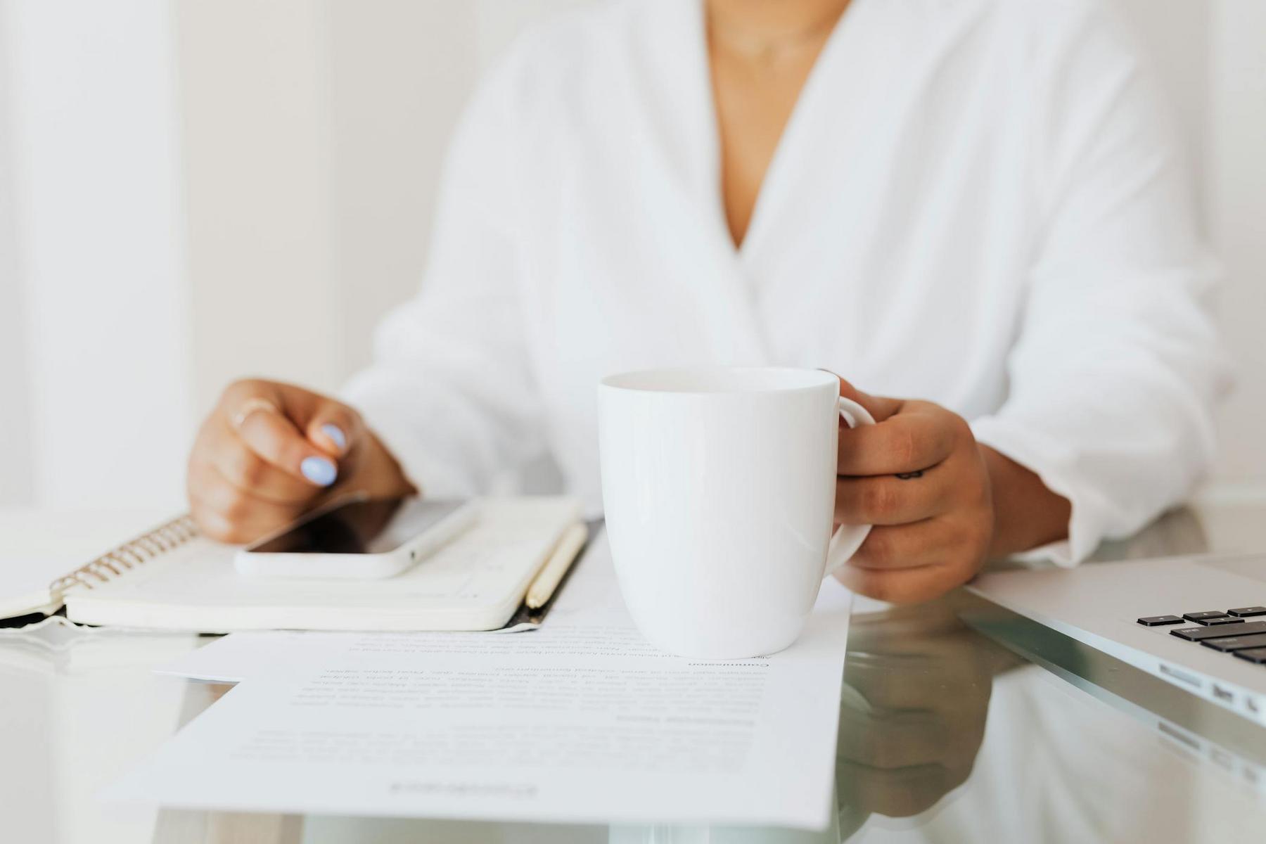 A person in a white shirt sits at a desk holding a white mug, with a notebook, smartphone, papers, and part of a laptop visible.