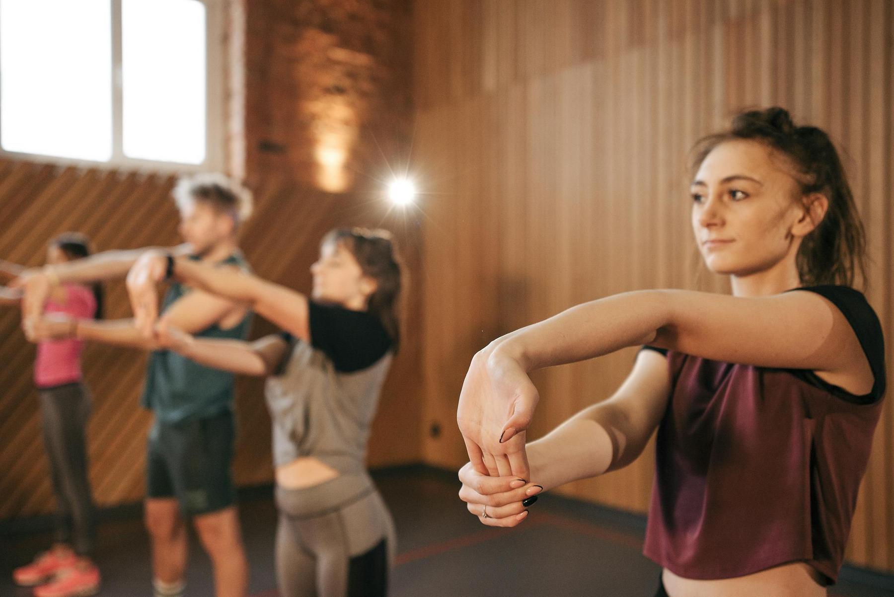 Four people stand in a row indoors, stretching their arms forward, with a wooden wall and window in the background.