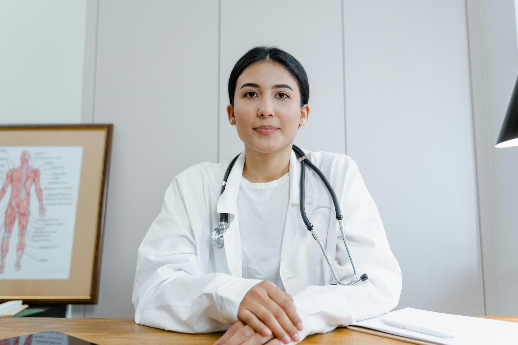A doctor wearing a white coat and stethoscope sits at a desk in an office with a medical diagram and papers visible in the background.