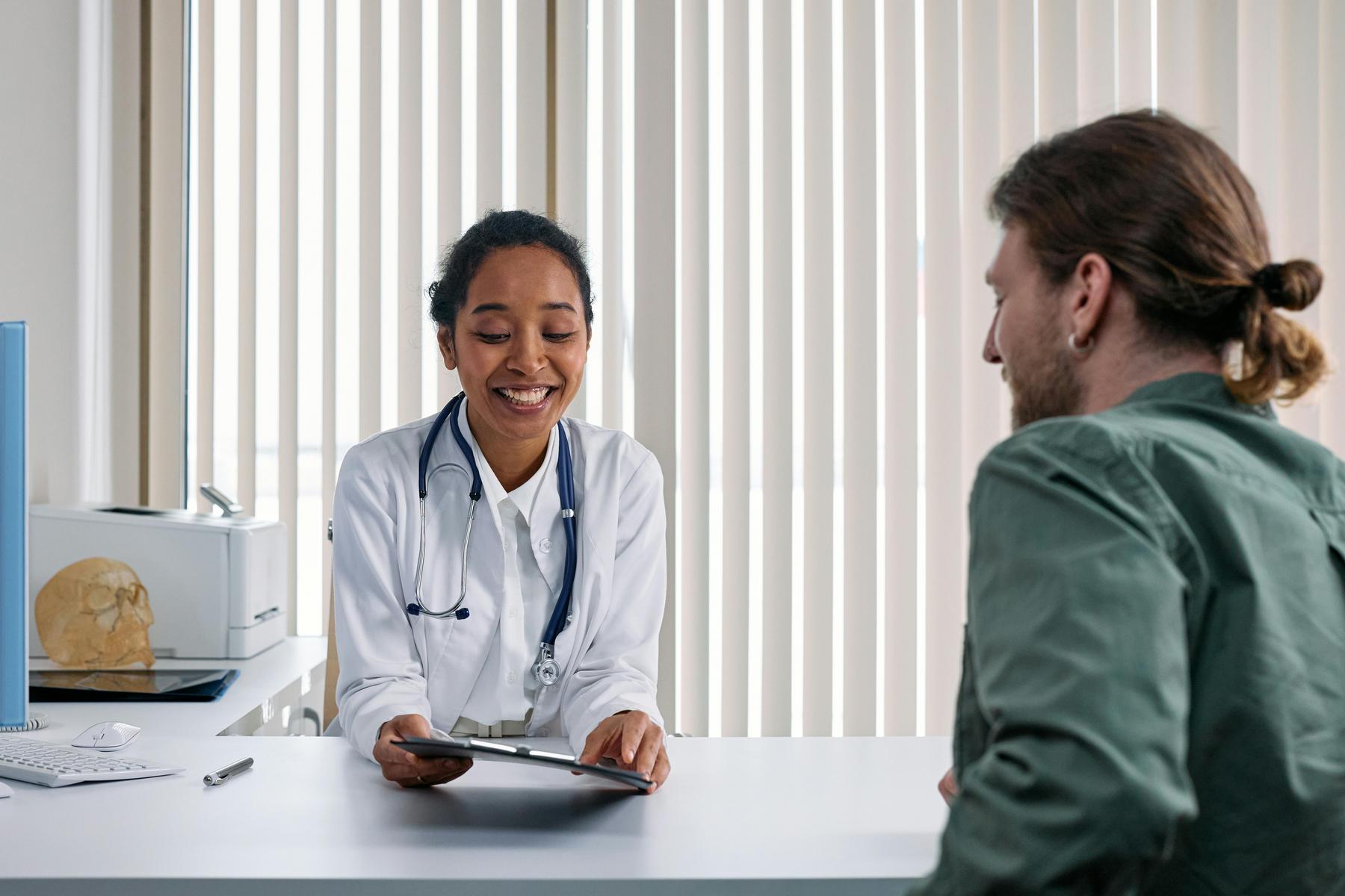 A doctor in a white coat shows a clipboard to a seated patient across a desk in a medical office with vertical blinds in the background.