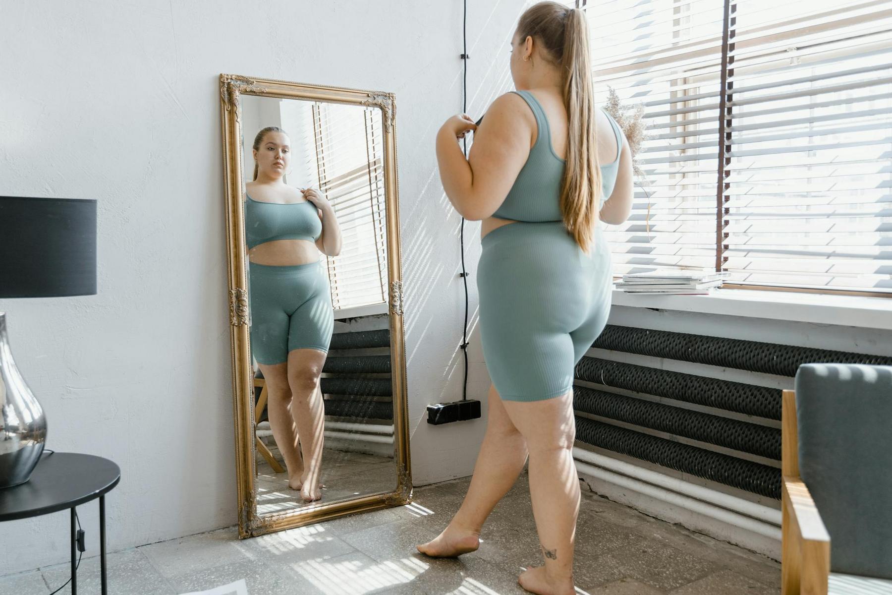A woman in light blue athletic wear stands barefoot in front of a tall mirror, looking at her reflection in a bright room with large windows.