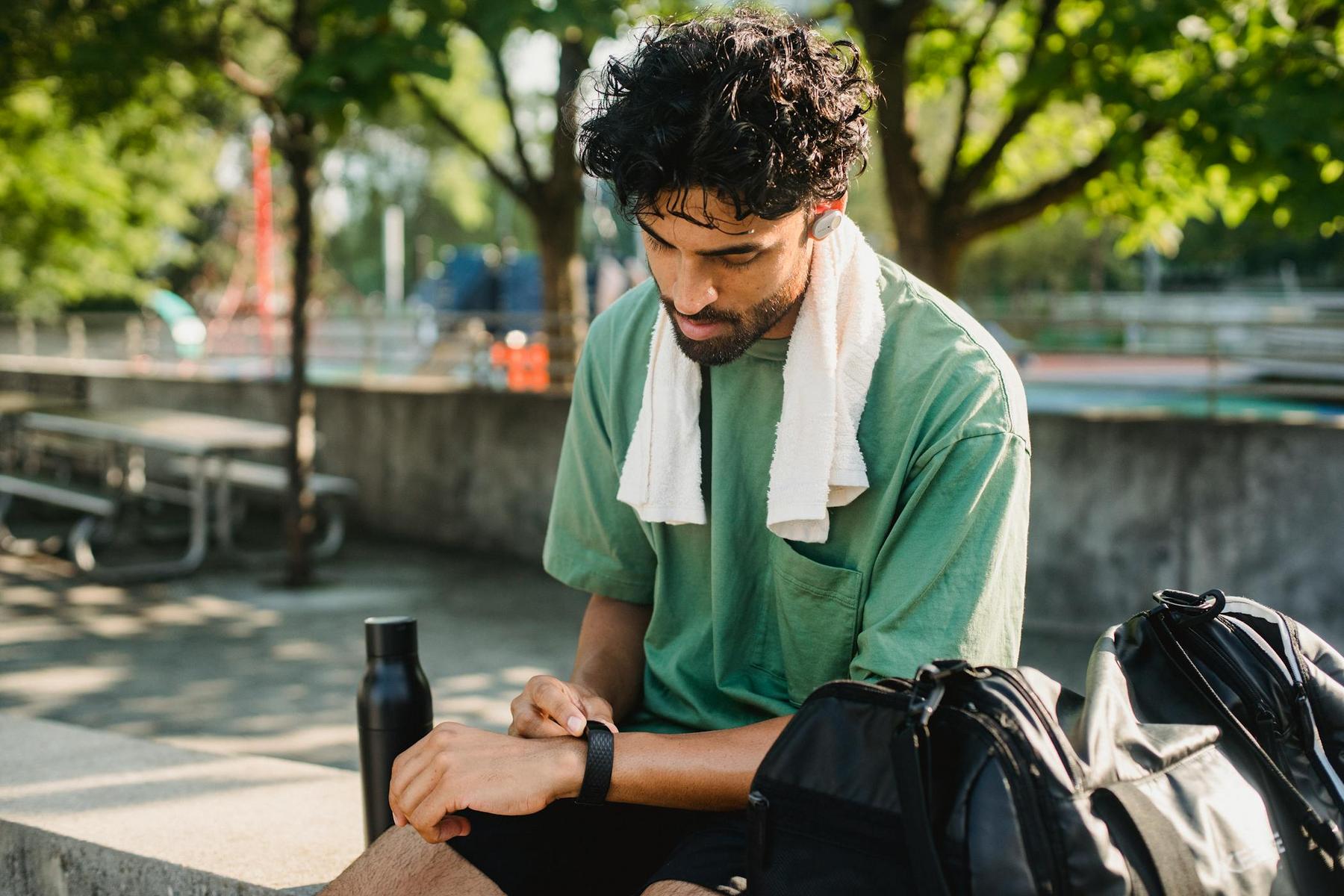 A man with a towel around his neck sits outdoors, looking at his smartwatch. A water bottle and a gym bag are next to him. Trees and a sports field are in the background.