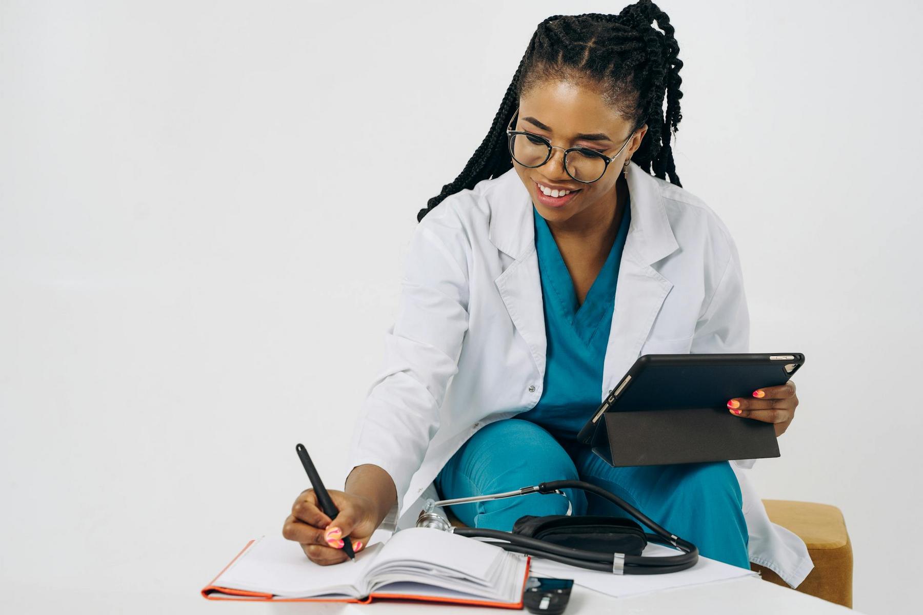 A healthcare professional in scrubs and a white coat writes in a notebook while holding a tablet, with a stethoscope and medical equipment nearby.