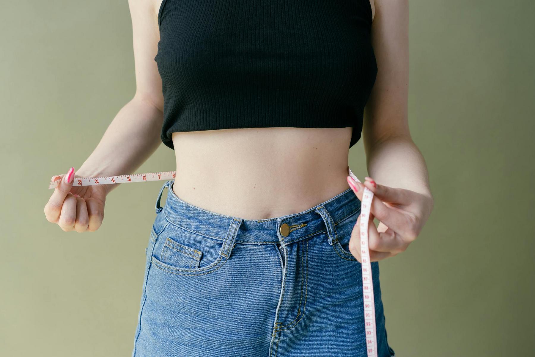 Person in a black crop top and jeans holds a measuring tape around their waist against a green background.