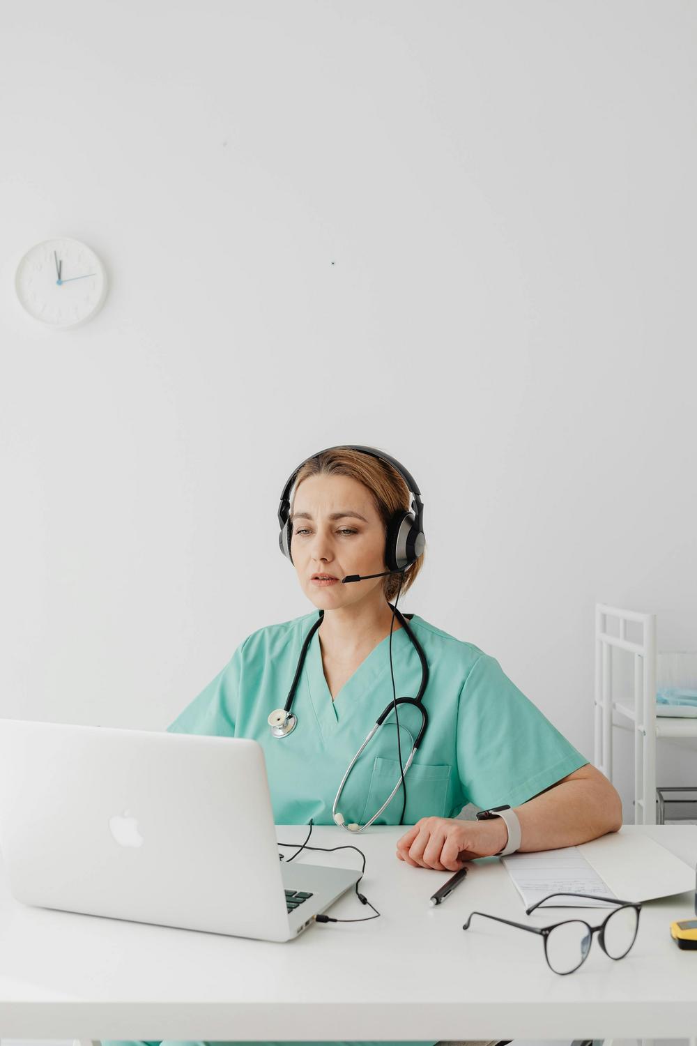 A healthcare worker in scrubs and a headset sits at a desk with a laptop, notepad, pen, and eyeglasses, conducting a virtual consultation.