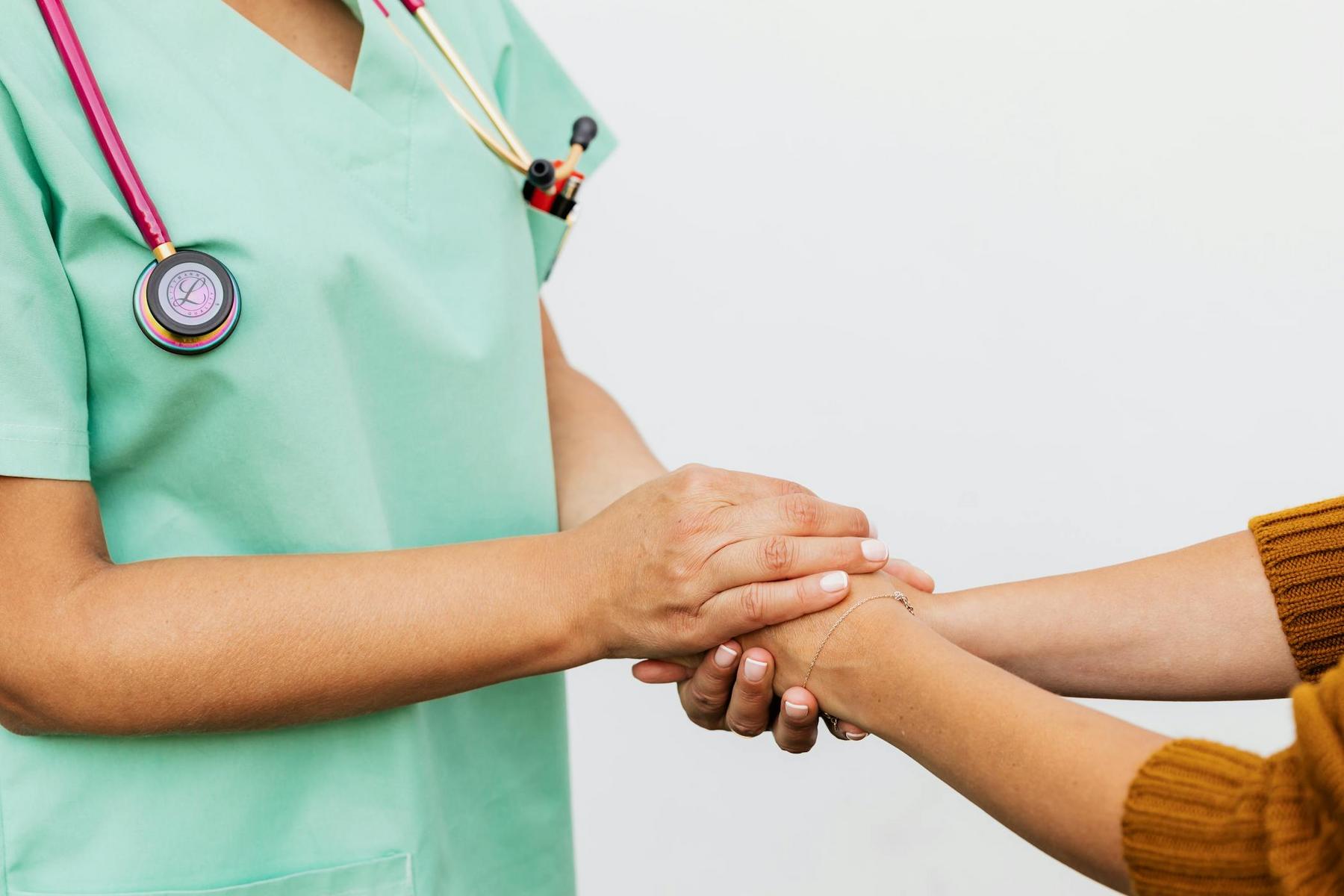 A healthcare professional in green scrubs holds the hand of a person in a brown sweater, offering support and reassurance.