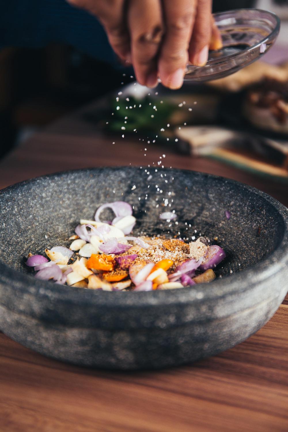 A hand sprinkles salt into a stone bowl containing sliced shallots, garlic, and spices on a wooden surface.