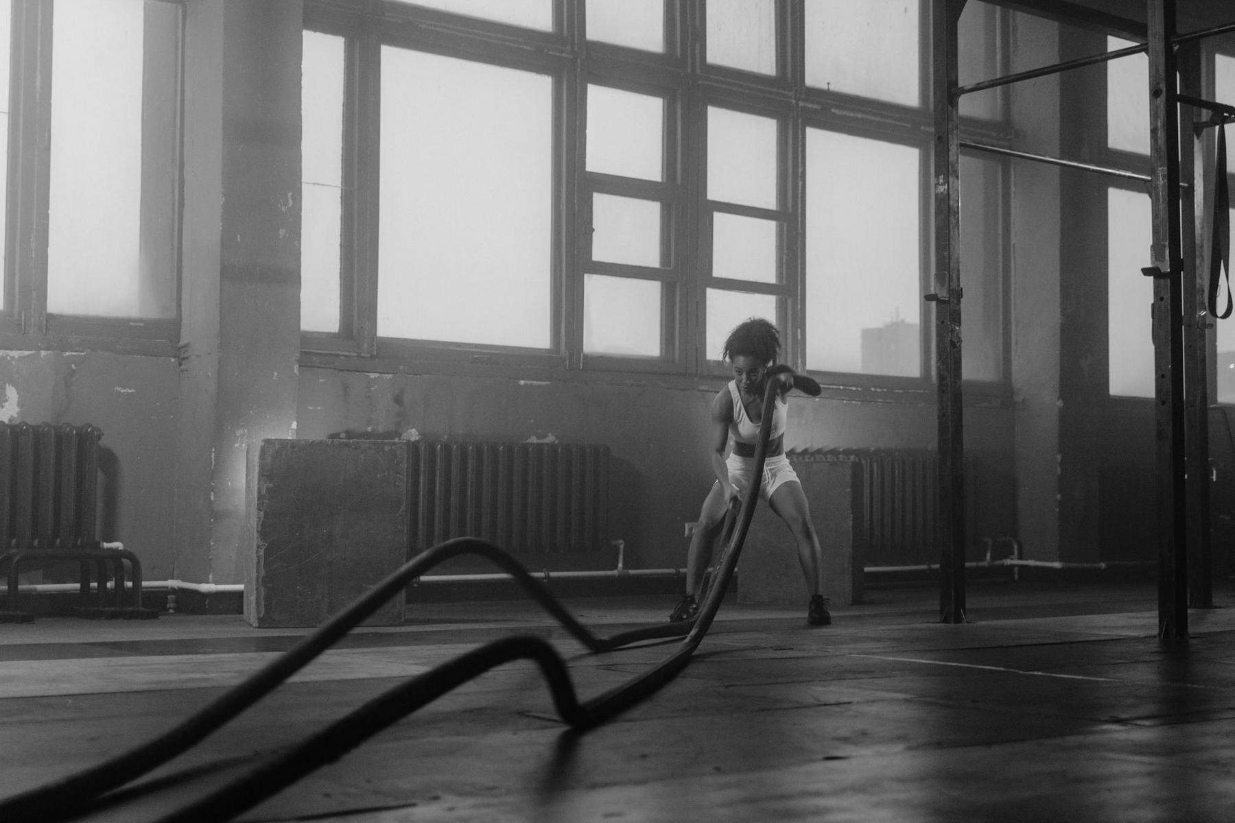 Person exercising with battle ropes in a gym, wearing athletic clothing, with large windows and industrial decor in the background.
