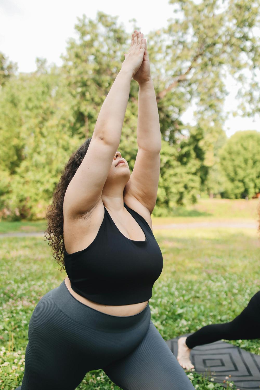 A person practicing yoga outdoors, standing on a mat with arms raised above their head, surrounded by green grass and trees.
