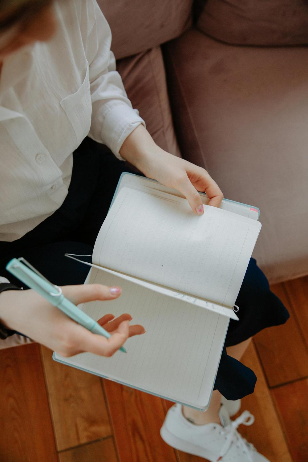 A person sits on a couch holding an open, blank notebook and a pen, with one hand resting on the pages and the other holding the pen.