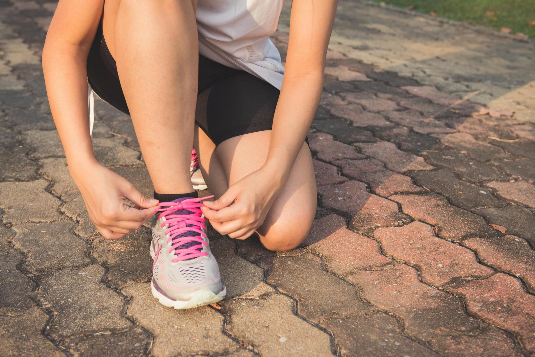 Person kneeling on a paved path, tying the pink shoelaces of a gray athletic shoe, wearing black shorts and a white top.
