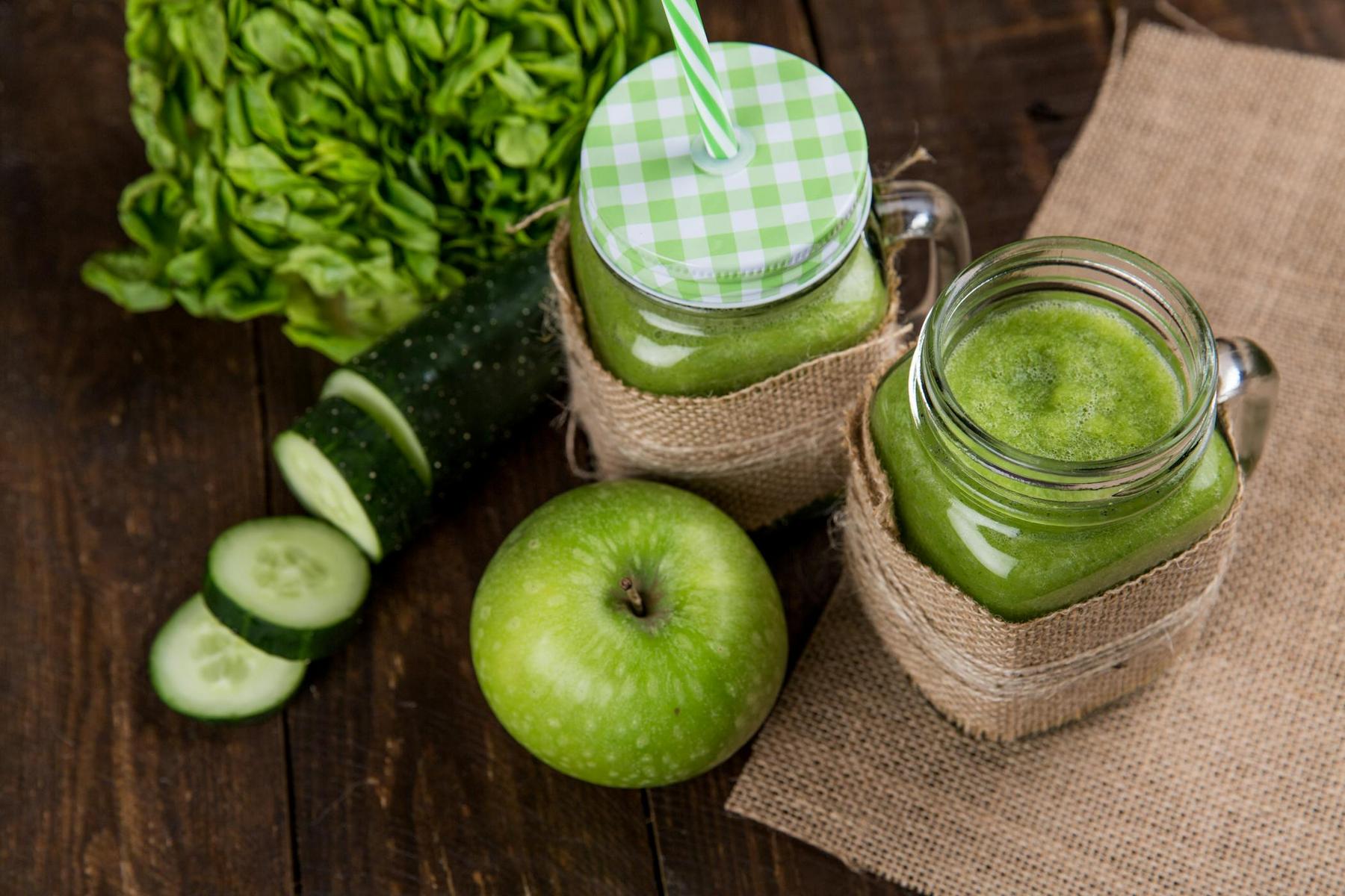 Two mason jars with green smoothies using striped straws, wrapped in burlap, near a green apple, sliced cucumber, and lettuce on a wooden table.