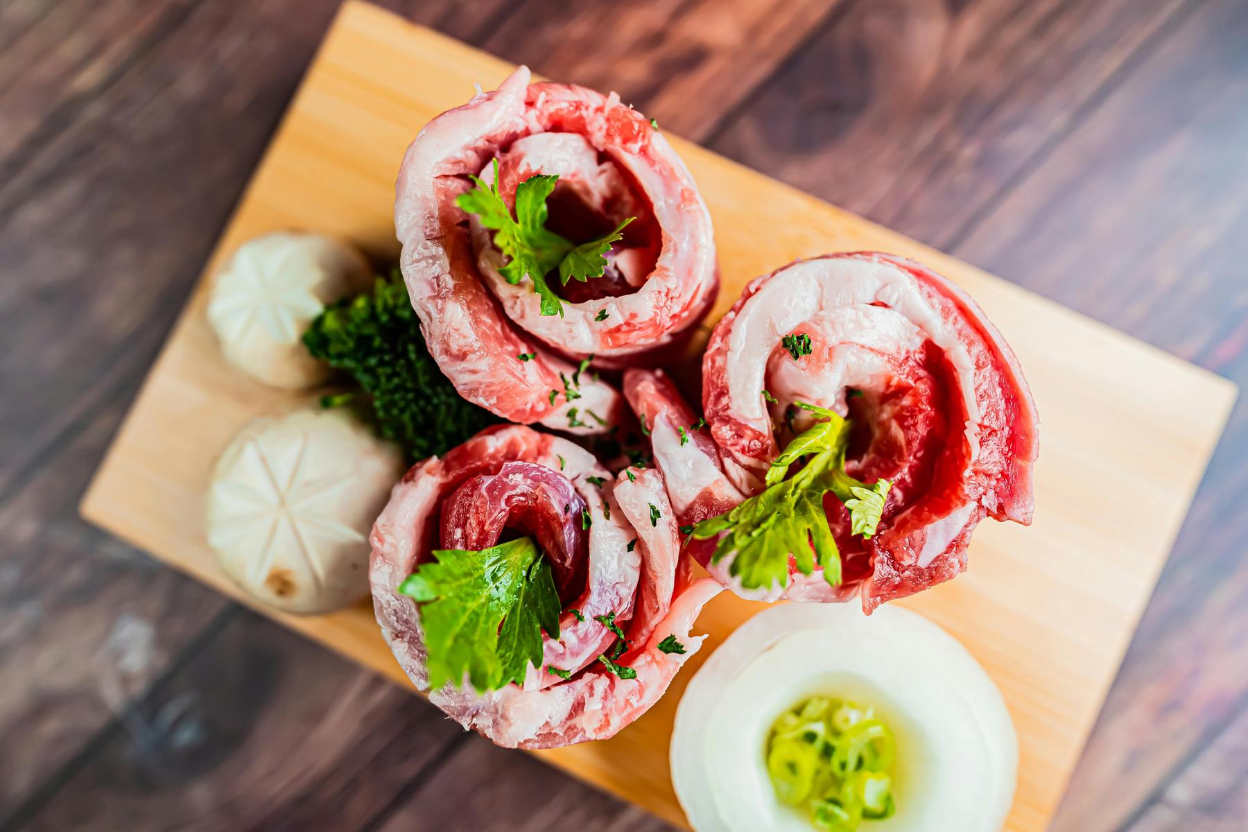 Raw meat rolls with parsley garnish on a wooden board, accompanied by mushrooms and a sliced onion.