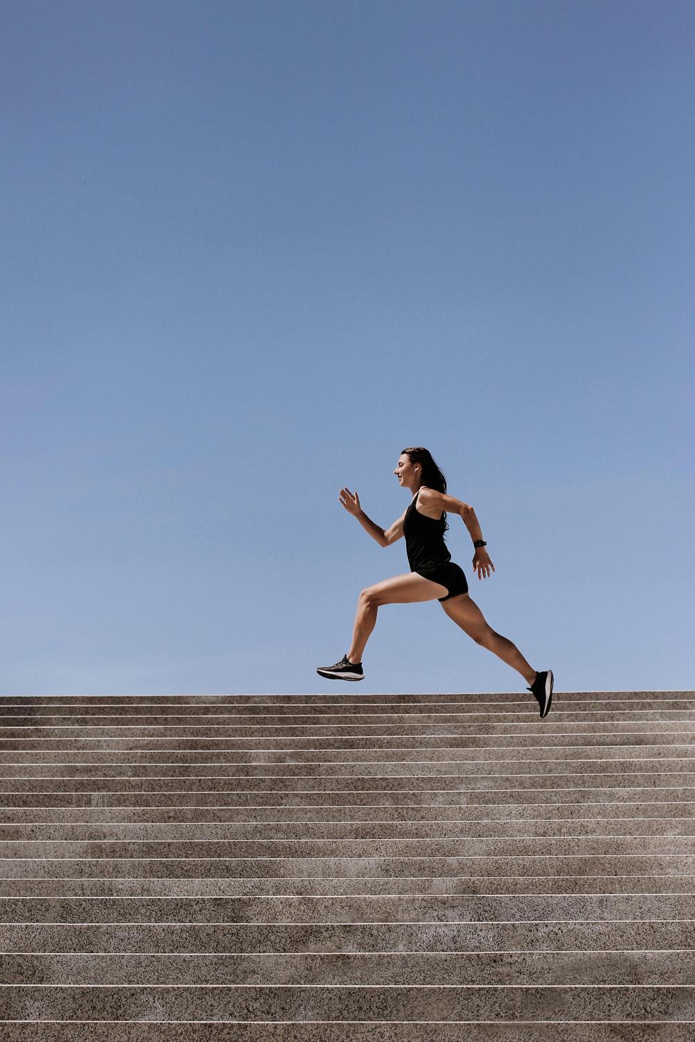 A woman in athletic clothing runs up outdoor concrete steps under a clear blue sky.