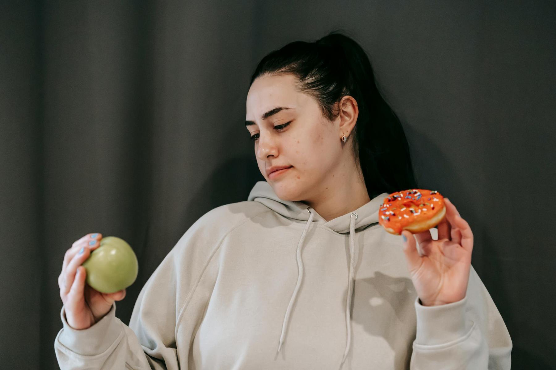 A person in a hoodie holds a green apple in one hand and a sprinkled donut in the other, appearing to contemplate the choice between them.