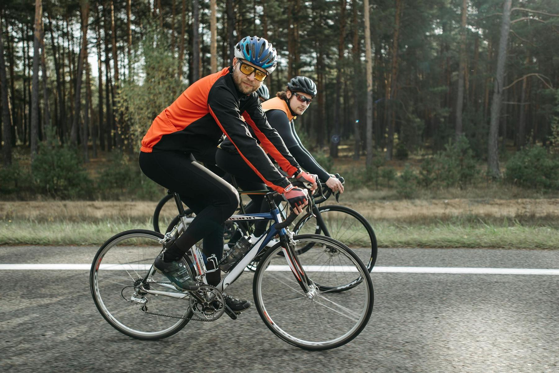 Two cyclists wearing helmets and sportswear ride road bikes on a paved road next to a forested area.