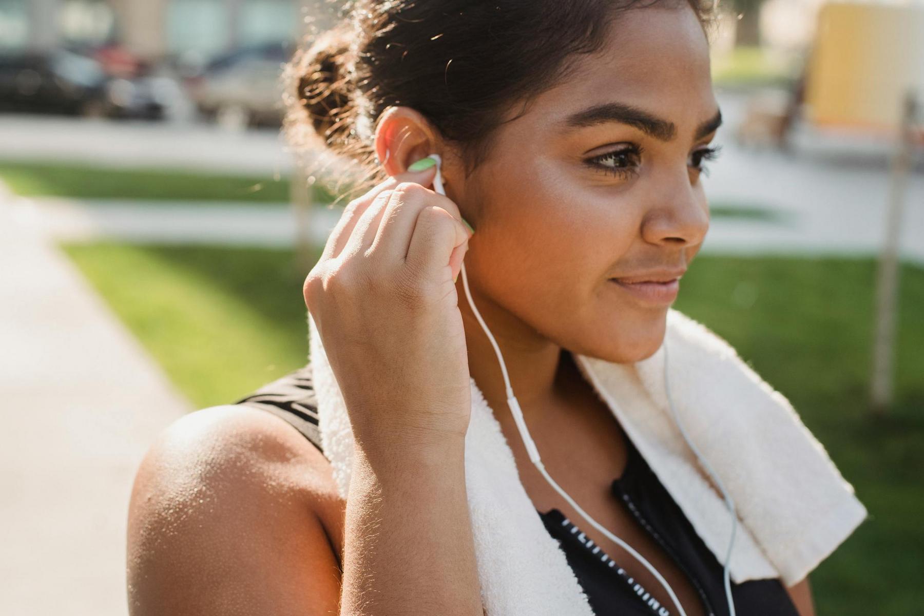 A woman with a towel around her neck adjusts her earphones outdoors, with a grassy area and blurred buildings in the background.