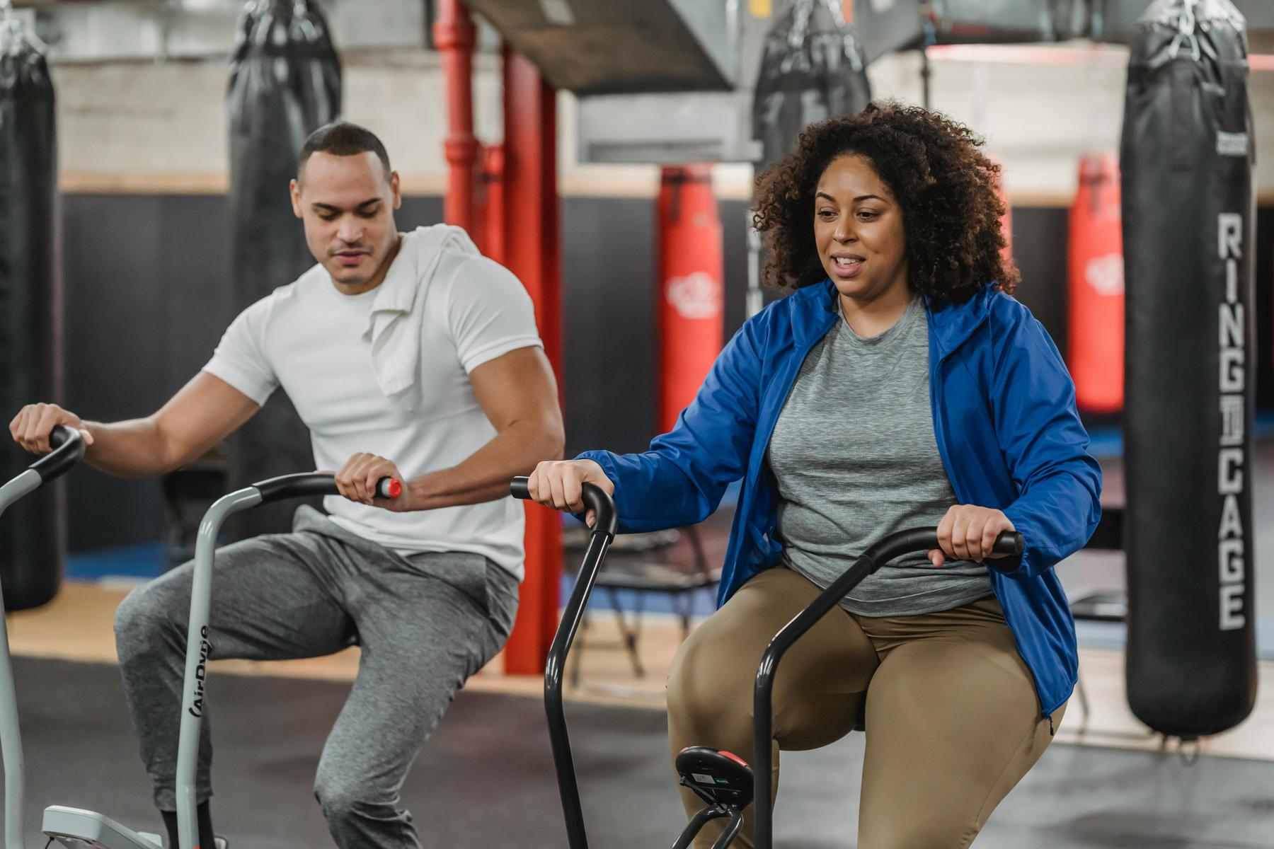 Two people exercise on stationary bikes in a gym with punching bags and red pillars in the background.