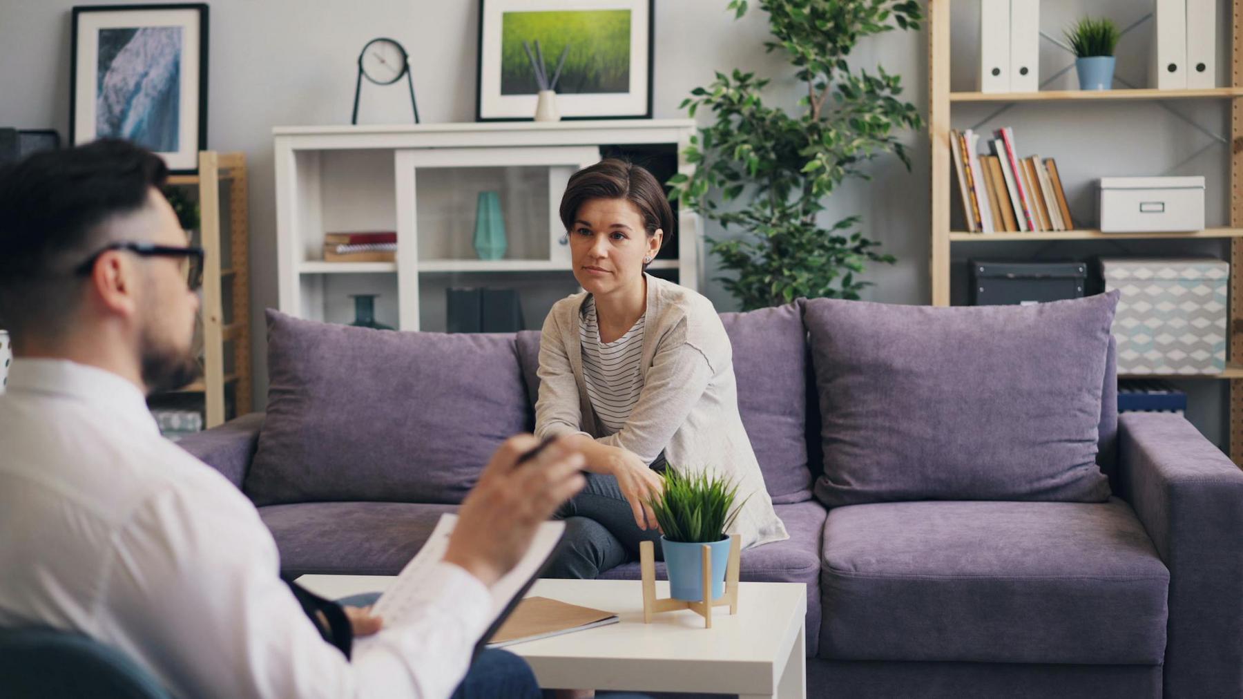 A woman sits on a purple couch, facing a man who appears to be a therapist. They are in a well-lit office with shelves, plants, and framed art in the background.