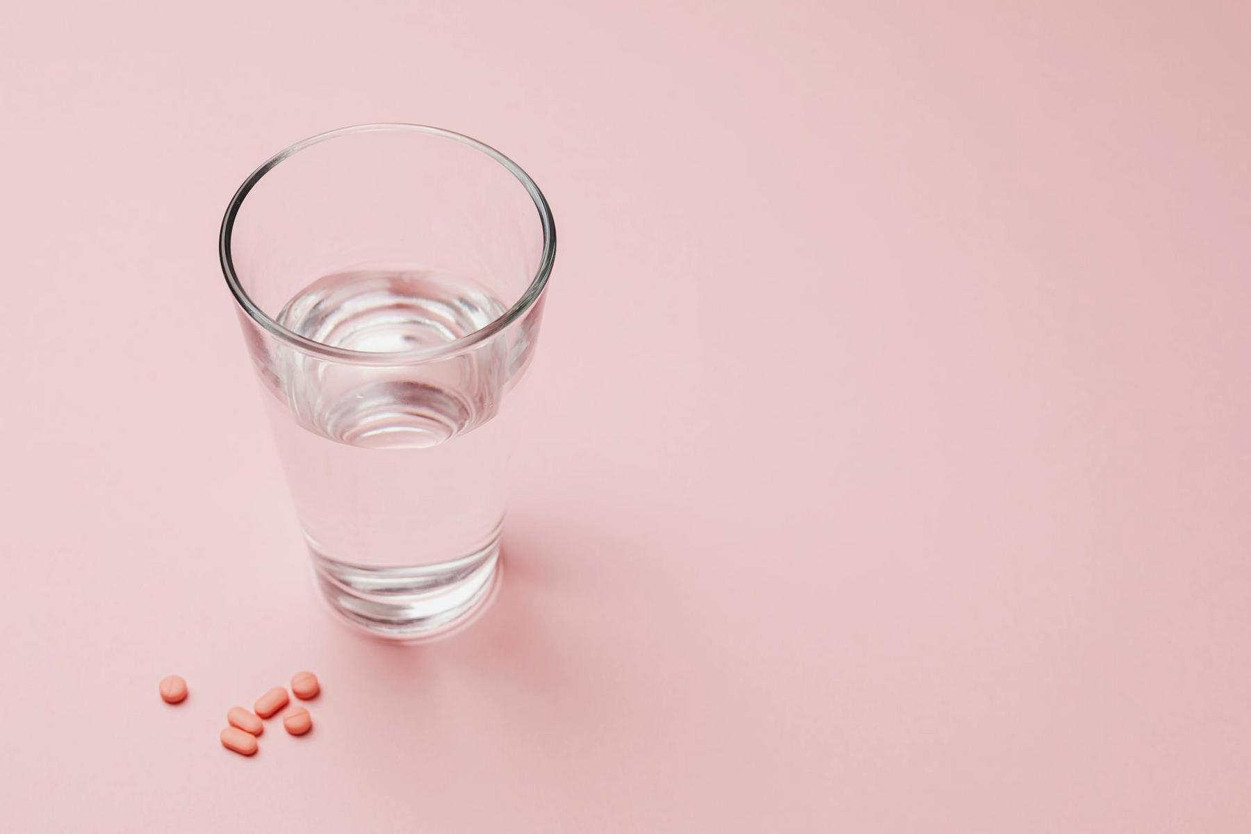 A glass of water sits on a pink surface next to a small cluster of red pills.