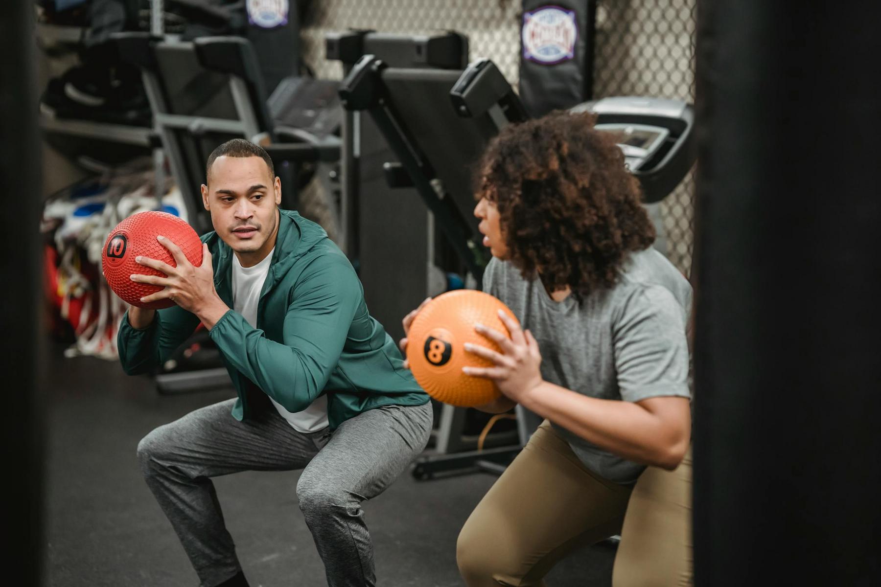 Two people perform squats in a gym, holding medicine balls and facing each other. Equipment is visible in the background.