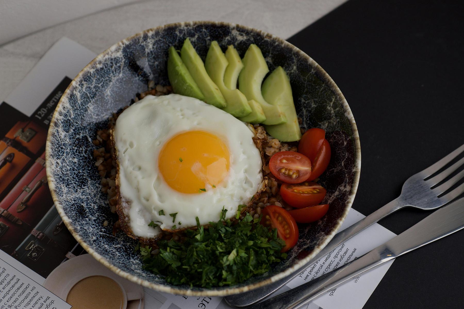 A bowl of rice topped with a sunny-side-up egg, sliced avocado, cherry tomatoes, and chopped parsley, next to a fork and knife on a magazine page.