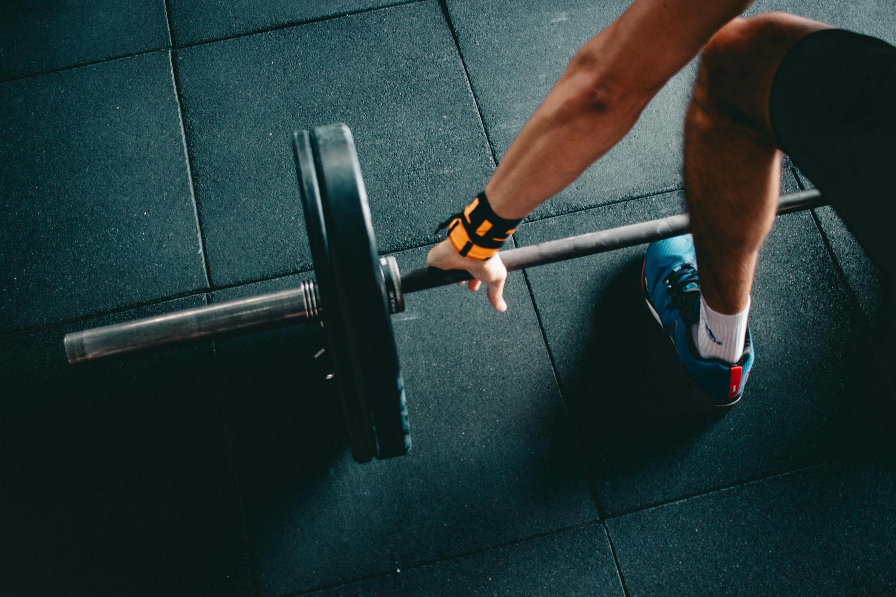 A person wearing a wrist strap and blue shoes prepares to lift a barbell with weight plates in a gym setting.